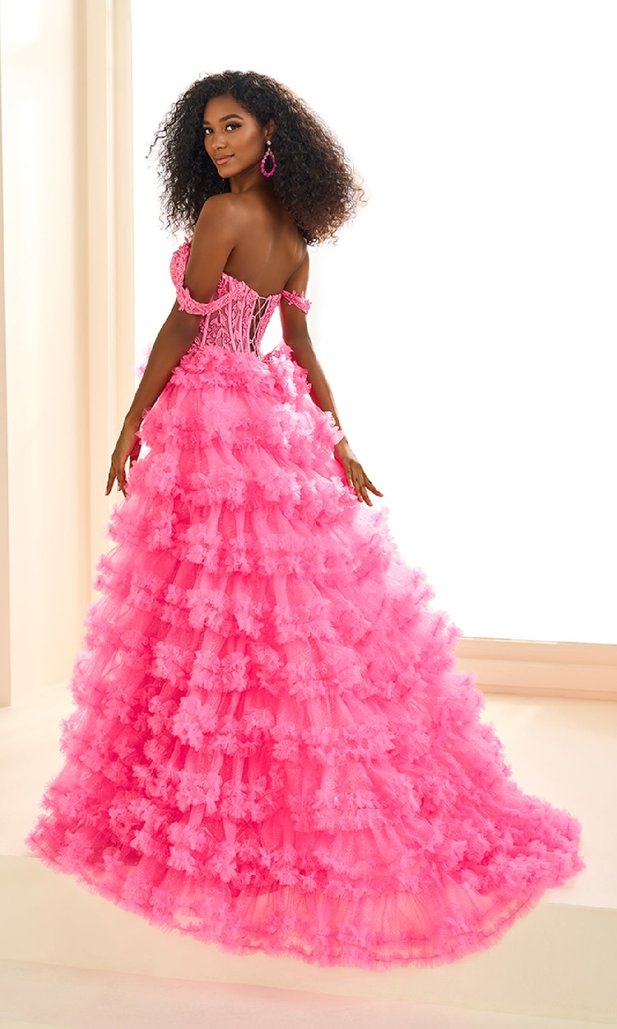Woman wearing the Ellie Wilde EW36201 long prom dress—bright pink, off-the-shoulder with a ruffled skirt and lace applique corset bodice—posing indoors by a large window.